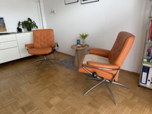 Zwei orangefarbene Sessel mit kleinem Tisch in der Praxis Michael Backes in Ratingen, ideal für vertrauliche Gespräche.