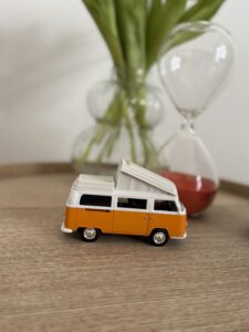 Kleines Modell eines orange-weißen VW Bulli vor einer Sanduhr, dekorativ platziert in der Praxis Michael Backes in Ratingen.